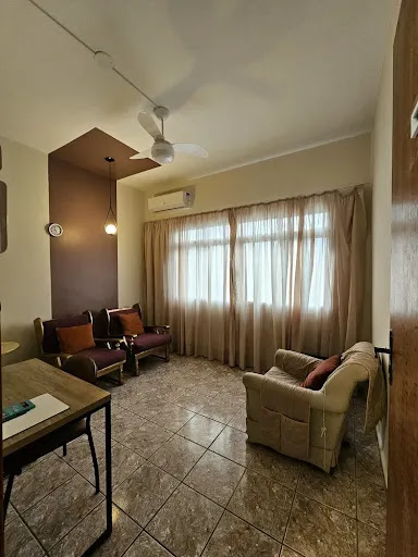 Sala de terapia clássica com poltronas confortáveis, escrivaninha de madeira, ar condicionado, ventilador de teto e cortinas leves.